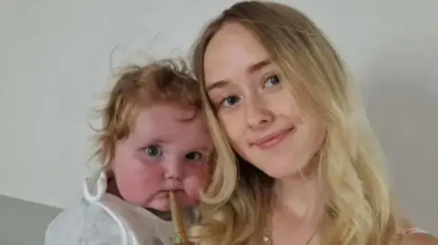 Family photo A woman with blond hair alongside a baby with blonde curly hair and a stick in his mouth, wearing a white top and bib