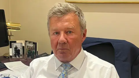 Eric Masterson sits at a desk in an office, wearing a white shirt and a blue floral-patterned tie. A dark blue jacket is draped over the chair behind him. The desk holds papers, documents, and two framed family photographs. A computer monitor and a brass desk lamp are visible in the background.