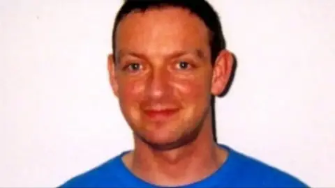 A man, with dark hair, smiling at the camera wearing a blue t shirt
