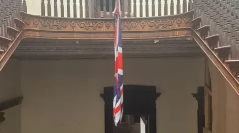 The Somme Association A brown ornate staircase with a union flag hanging above an ornate door frame with cream walls.