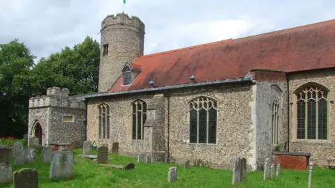Simon Knott Church is shown with porch to the side. It has a round town. Gravestones sit in the foreground