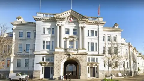 Google The outside of Kent County Council county hall. The building is three storey's high and made of brick. Lots of the brick is painted white. At the centre of the building is an archway. 