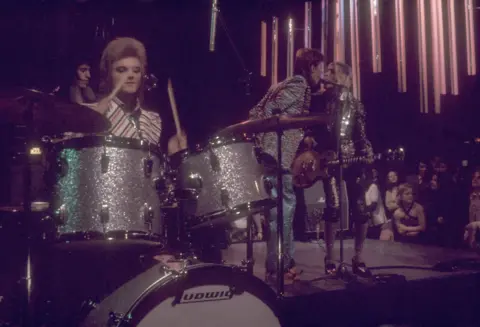 Watal Asanuma/Shinko Music/Getty Images An archive image of the Spiders from Mars on Top of the Pops in 1973. A drummer playing a sparkly silver drum kit sits front left of the picture. David Bowie is stood in the background singing into a microphone alongside Mick Ronson. They are both wearing animal print outfits.