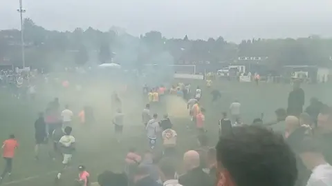 @instructormannn/Andy C An image taken from the stands of a football ground, showing dozens of people on the pitch. The view is obscured by clouds of green and yellow smoke drifting across the pitch.