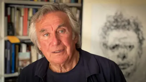 Jeff Towns looking at the camera with a walls of books on the shelf behind him and black and white portrait of Dylan Thomas