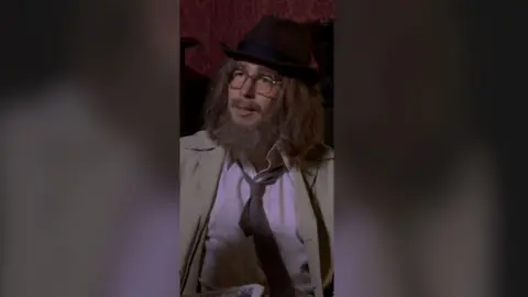 A man in costume - with a hat, glasses, long hair and a long beard. He wears a tie loosely.