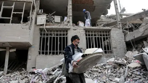 WANA/via Reuters A man walks amid rubble from a building destroyed by a strike in Tehran, Iran (12 March 2026)