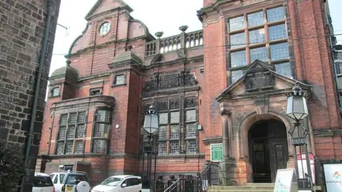 A view looking up to a red brick building which has dark rectangle-shaped windows and stone steps leading into it. There are some signs outside and several parked cars. 