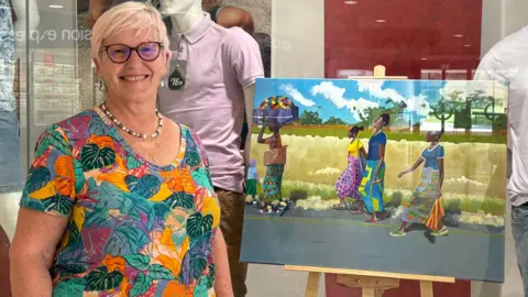 Gloucester Contemporary Artists Gilly Hill. She has short fair hair and glasses. She is stood next to her painting which is on display in a shop window.