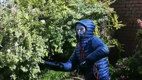 A person in a blue anorak, with the hood up and a mask over the face, begins to throw an unknown package at a green bush.