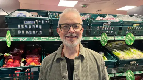A slim man with cropped white hair and a matching beard smiles directly at the camera. He is wearing a green shirt and dark-framed glasses. He stands in front of rows of bags and punnets of fruit and vegetables, such as nectarines, cranberries and leeks, which are all priced  at 20p with big green labels on the shelves.