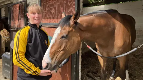 Rachel Carton, a woman with blonde hair styled with a fringe, poses for a photo with Spirit Dancer the former racehorse. The horse is standing in a stable, with its muzzle buried in Carton's hand. She is wearing a black tracksuit top with yellow sleeves.