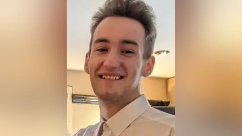 A closely cropped photo of a young man smiling. He has stubble and is wearing a white shirt. 