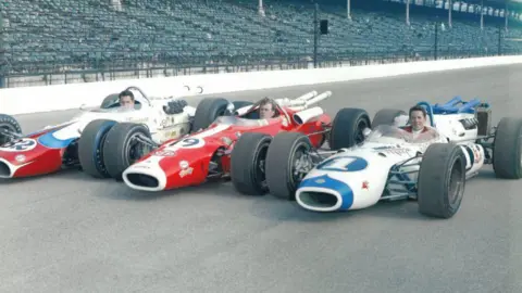 Gildings Auctioneers Mike Underwood sitting in the 1966 Indy 500 Lotus (central car) alongside drivers Mario Andretti (left), and George Snider (right). All three cars are on the track.