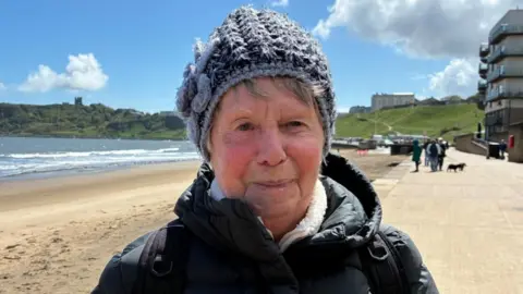 An older woman with grey hair looks at the camera. She wears a grey knitted hat and a black padded waterproof jacket. Behind her, a view of a beach and sea can be seen. 