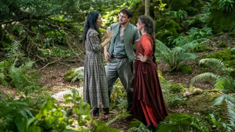 Universal Pictures [L-R] Behind-the-scenes image showing director Chloé Zhao on the set of Hamnet with Paul Mescal and Jessie Buckley
They are standing in the middle of a lush green forest.
Chloe with long black hair in long checked grey dress
Paul in moss green top and trousers
Jessie in dark red dress