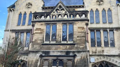 A close up of a church building with stained glass windows. Some of the brick is bright and Cotswold yellow while other parts are really black. On the left hand side ivy is crawling up the building and is on one of the windows.