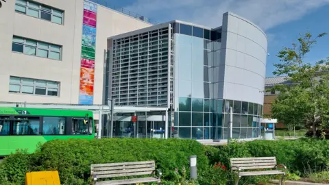 Owen Ward/BBC A hospital building with a green hedge and benches in the foreground. A green bus is also visible. There is blue sky and some cloud above.