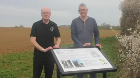 Northamptonshire Battlefields Society Graham Evans in a black shirt standing beside Simon Marsh in a grey long sleeved top standing on the grassy edge of a ploughed field. In front of them is an information board about the Battle of Edgcote. 