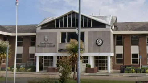 A picture taken outside Castle Point Borough Council's offices. The building has the council's name on the front of it and a clock. There are white columns by the entrance door and it has a triangular roof. There is a flag pole in front of the building.