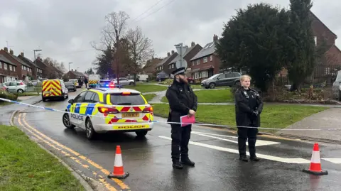 Two police officers stood behind the cordon closing off Hazel Avenue, Guildford, between the junctions with Lime Grove and Willow Way. Several police cars and a van can be seen in the background further up the street.