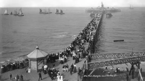 Getty Images A black and white photograph of Margate Pier taken between 1890-1910. Crowds of people in contemporary Victorian clothes line the wooden and cast-iron structure. Sail boats can be seen in the distance on a flat sea. 