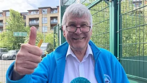 A man with grey hair and glasses, wearing a pale shirt under a blue fleece, gives the camera a thumbs-up.