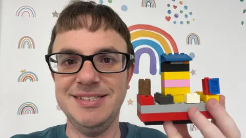 A selfie of a man wearing black-rimmed glasses holding a multi-coloured Lego construction in his left hand.