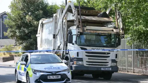 A white police car parked next to a white bin lorry in a street with blue and white police tape in front of them
