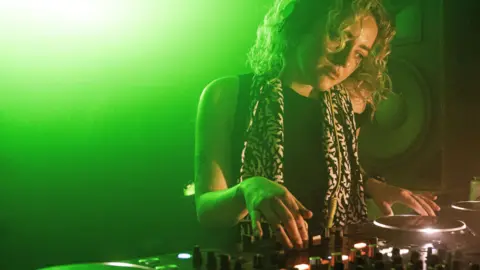 Jacinta Owen Jacinta has blonde, curly medium-length hair. She wears a black top and a black and white scarf. She stands in front of DJ decks
