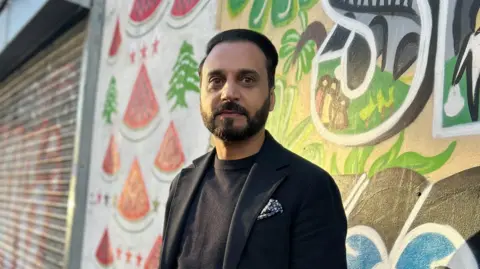 Abdul Malik wearing a black jumper and suit jacket, photographed outside against a background of street art.