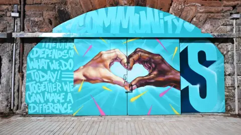 A large artwork on a railway arch. The painting depicts two hands creating a heart, one with dark brown skin and one with white skin. Writing on the painting reads "The future depends on what we do today! Together we can make a difference."