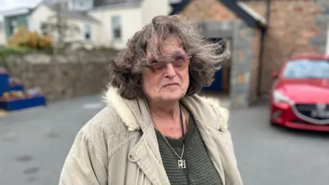 Corrine Joy wearing a dark top and a cream coloured coat. She has dark brown/grey hair.