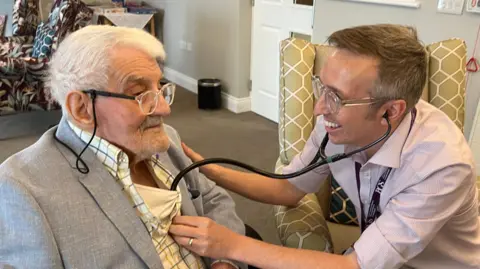 Clifford Craske is on the left and is having his heart checked by Matthew Butler, who is on the right and is using a stethoscope. Craske has short white hair and is wearing clear glasses, a white vest, white checked shirt and a grey suit jacket. Butler has short brown hair and is wearing clear framed glasses, a pink shirt and black lanyard.