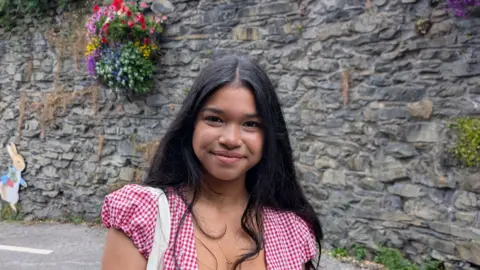 Saba Nur has long dark hair and dark eyes. She is standing in front of a stone wall with a colourful flower basket and a picture of Peter Rabbit. She is wearing a red and white gingham blouse.