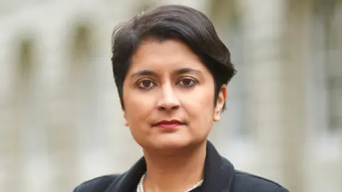 Shami Chakrabarti is looking at the camera and has short brown hair and brown eyes, and is wearing a black coat. The background behind her is blurred.