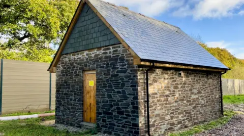 CEC A stone barn with a slate roof and black guttering and a brown wooden door set next to a brown fence and with blue sky and clouds, some trees and grass around it