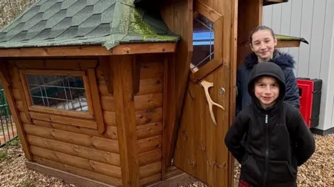 Sarah McCaffrey A young boy in a black hoodie with the hood up stands to the right of the image, a young girl with a dark blue coat and faux-fur collar stands behind him. They are standing next to a small wooden house.