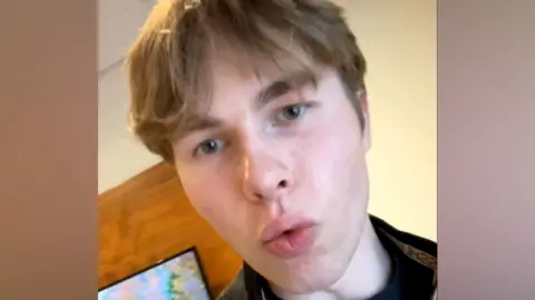 A selfie of Johannes Natland. He has short blonde hair and dark eyebrows. His lips are pursed as though talking. He is wearing a black top.