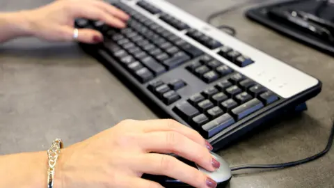 BBC A woman holding a computer mouse and hovering her hand over a keyboard on a grey desk.
