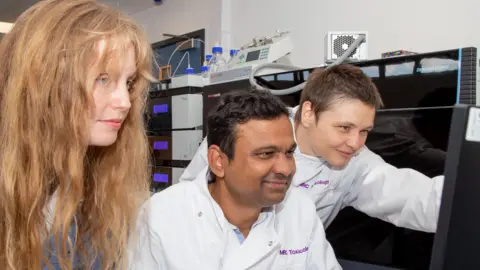 MRC Toxicology Unit The three researchers are looking at a computer screen while in a lab. Sonja Blasche has her arm outstretched to the screen and has short brown hair. All three are wearing white lab coats with the purple branding for MRC Toxicology Unit.