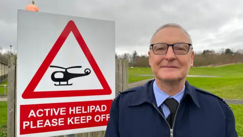 Tim is looking into the camera, you can just about see the helipad behind him. To his right is a red and white sign with a picture of a helicopter on it, the sign reads 'active helipad please keep off.'