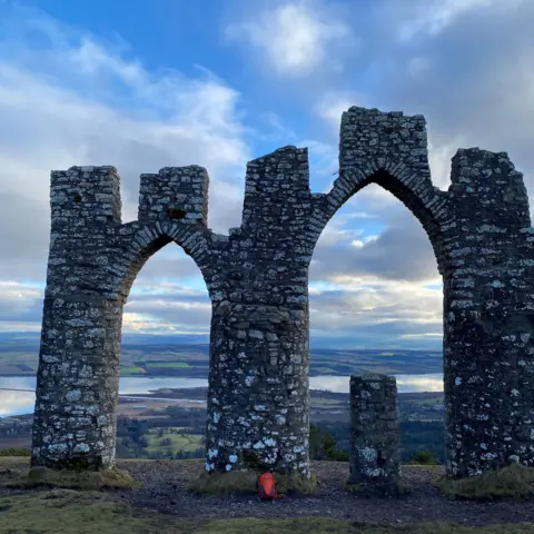 Robert Davidson The Cnoc Fyrish arched stone monument which is a mock ruin, called a folly, with water and hills in the distance under a cloudy sky.