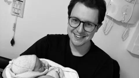 A black and white photograph of Daniel holding baby George in hospital who is wearing a beanie hat and wrapped in towels