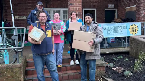 Glenroy Bell Four people standing outside a building, on steps, holding boxes. There is a sign to the right. They are all looking at the camera and smiling. 