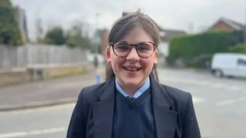 The head and shoulders of Amalie are visible in the foreground. She is smiling and looking at the camera. Amalie is wearing a navy v-neck jumper with a dark blue tie and light blue shirt underneath. She has a dark blue blazer on top. Amalie has dark brown medium length hair tucked over her shoulders and a fringe and is wearing tortoiseshell coloured round framed glasses. In the background is a T-junction in a residential area.