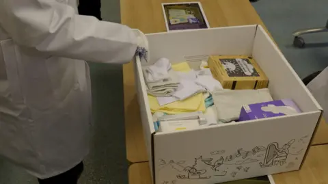 Welsh government The baby bundle on a wooden desk being packed up with items like clothing blankets, bibs, muslin cloths, a thermometer, a bilingual book and a playmat.