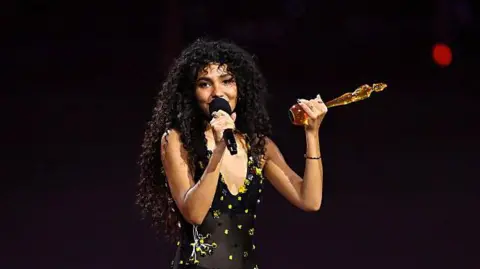 Olivia Dean, a woman wearing a black dress with yellow dotted patterns, speaking into a black microphone and holding a golden trophy.