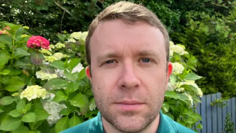 Peter Reynolds is looking into the camera. His brown hair is short. He's wearing a green top. Flowers and bushes are behind him and 