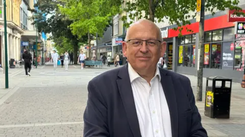 Chris Sutton, a man wearing a navy suit jacket and white shirt, smiles at the camera standing on Newport's main shopping street